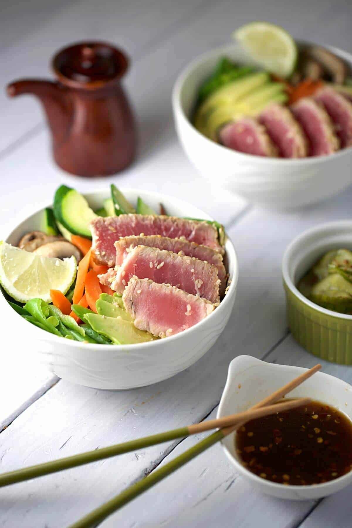Sliced sesame-crusted ahi tuna served over bowls with vegetables, lime, pickled cucumbers, and dipping sauce.