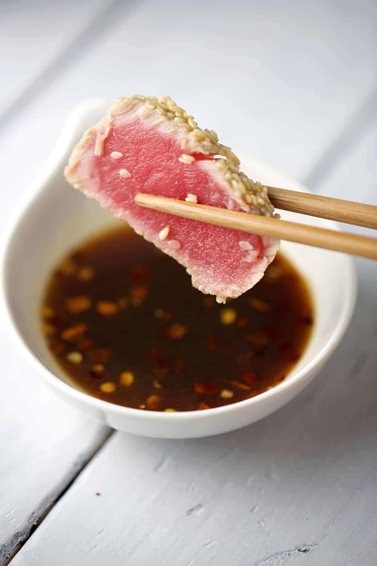 Chopsticks dipping a sesame-crusted ahi tuna slice with rare pink center into soy ginger lime sauce.