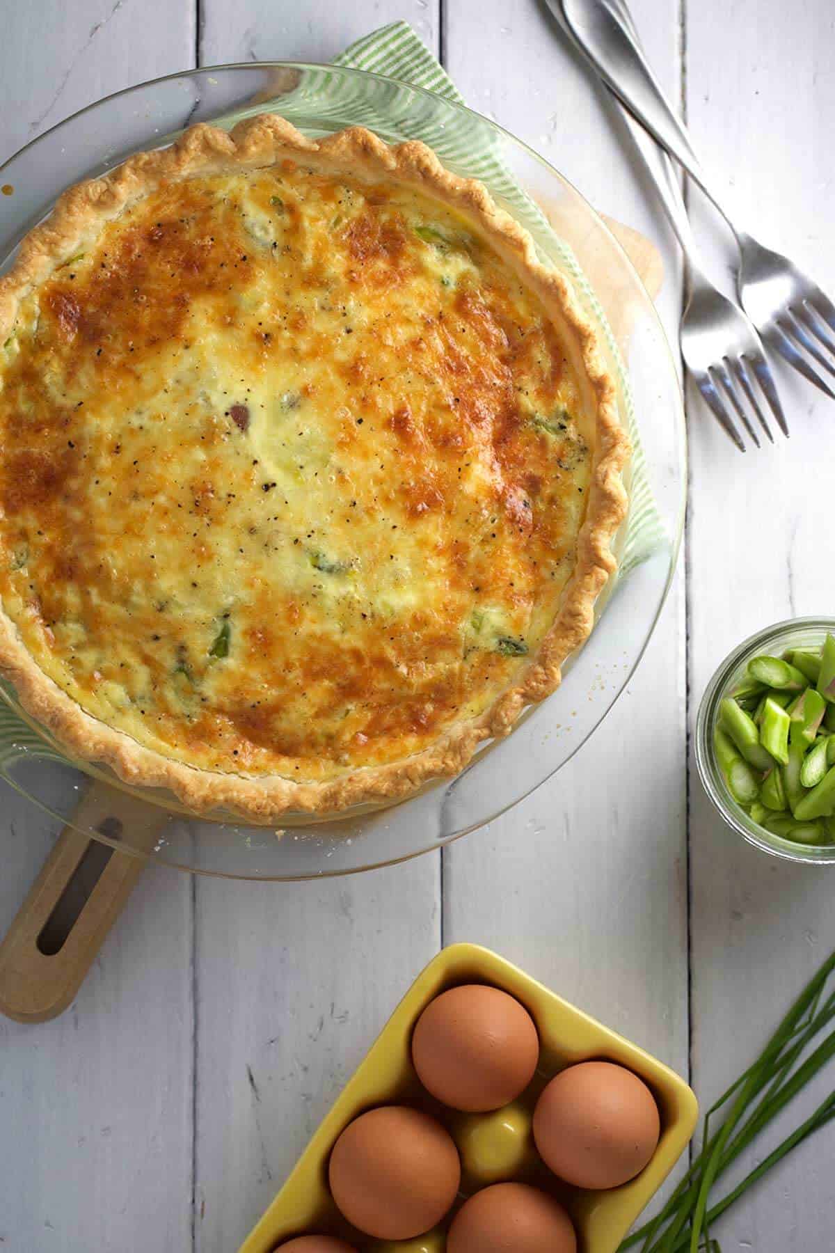 Quiche in a baking dish with eggs and asparagus next to it.