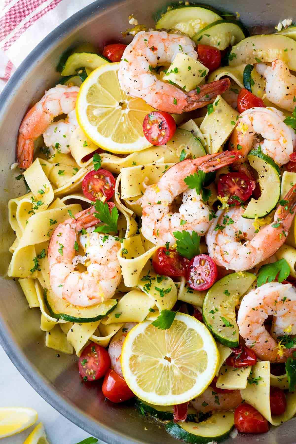 Pappardelle pasta with cooked shrimp, sliced zucchini, halved cherry tomatoes, and lemon slices in a skillet.