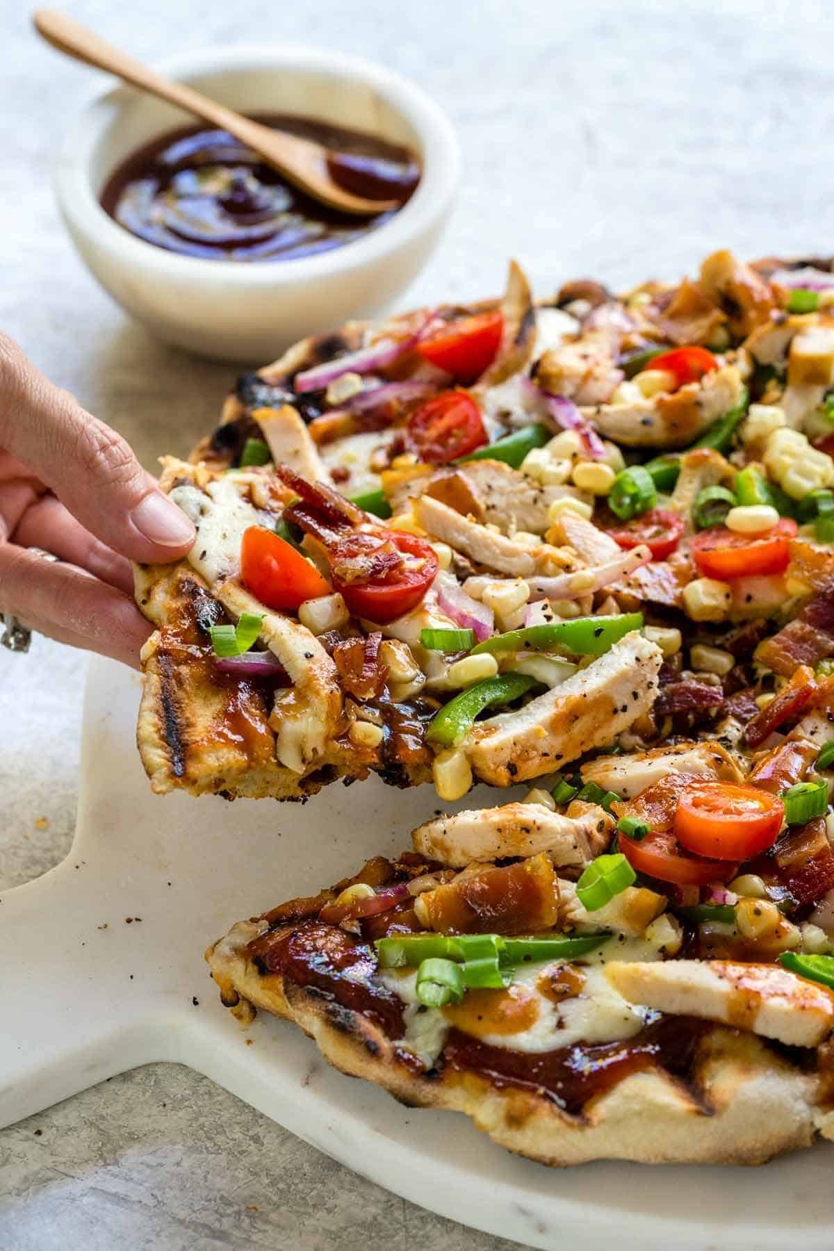 A hand lifting a slice of grilled BBQ chicken pizza topped with sliced chicken, tomatoes, corn, and peppers.