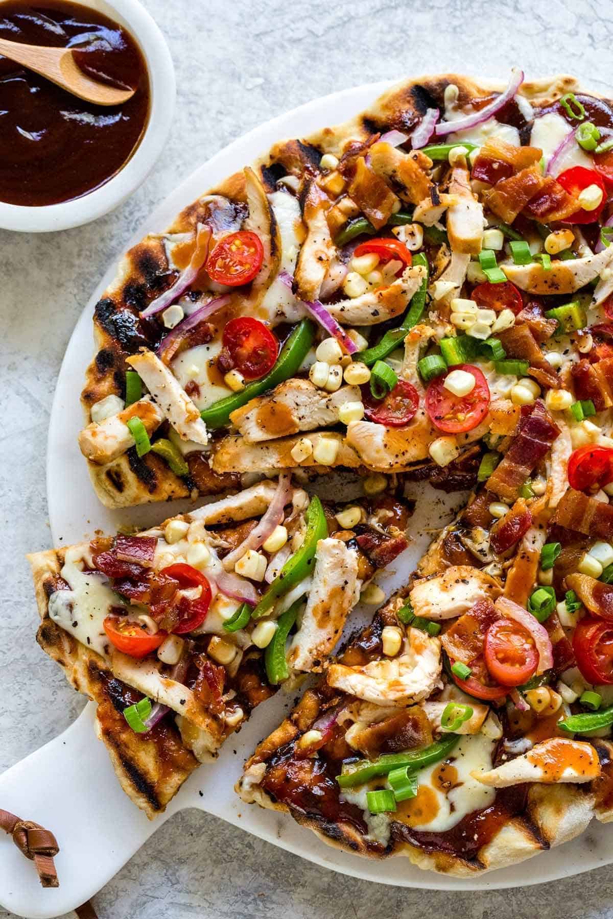 Grilled BBQ chicken pizza sliced on a serving board, topped with chicken strips, corn, peppers, bacon, and tomatoes.