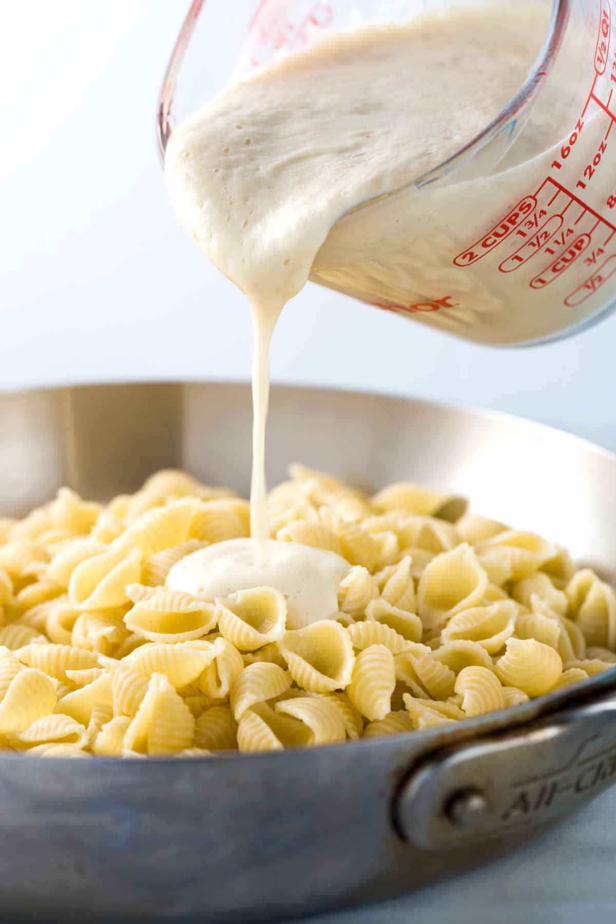 Measuring cup pouring cauliflower alfredo sauce over a pan of pasta noodles.
