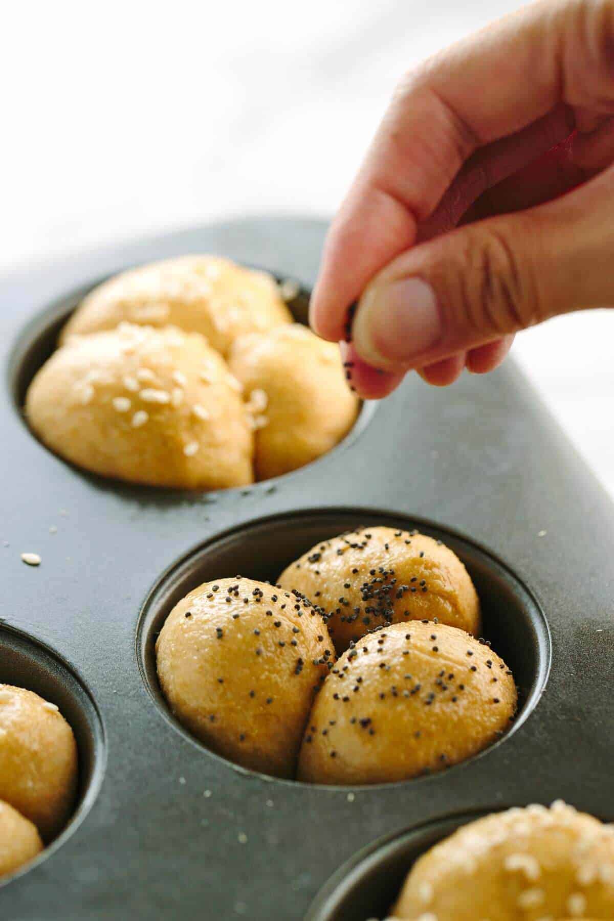 fingers sprinkling poppy seeds over dough balls in a muffin tin