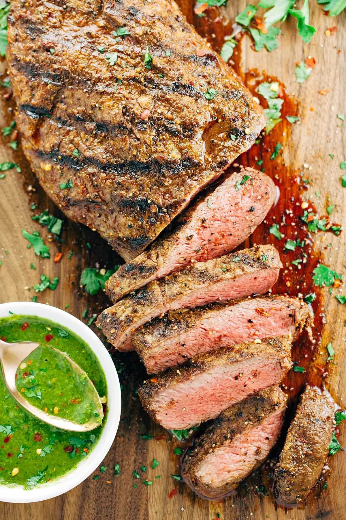 Sliced grilled steak cooked medium-rare on a wooden cutting board served with a bowl of green sauce.