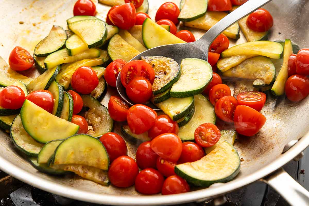 Sliced zucchini and cherry tomatoes sautéing in a stainless steel skillet with a metal spoon.