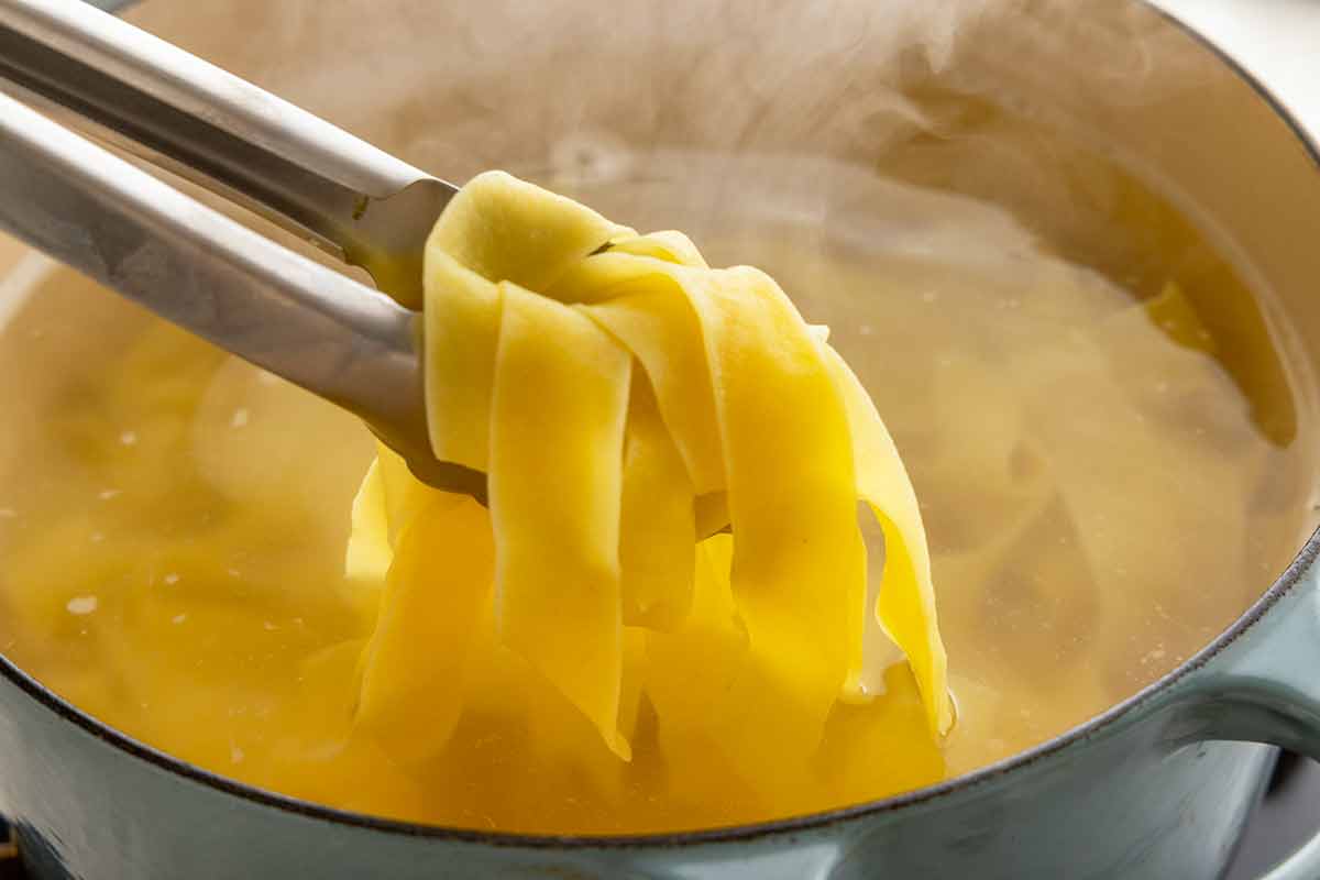 Metal tongs lifting wide pappardelle pasta ribbons from a pot of boiling water.