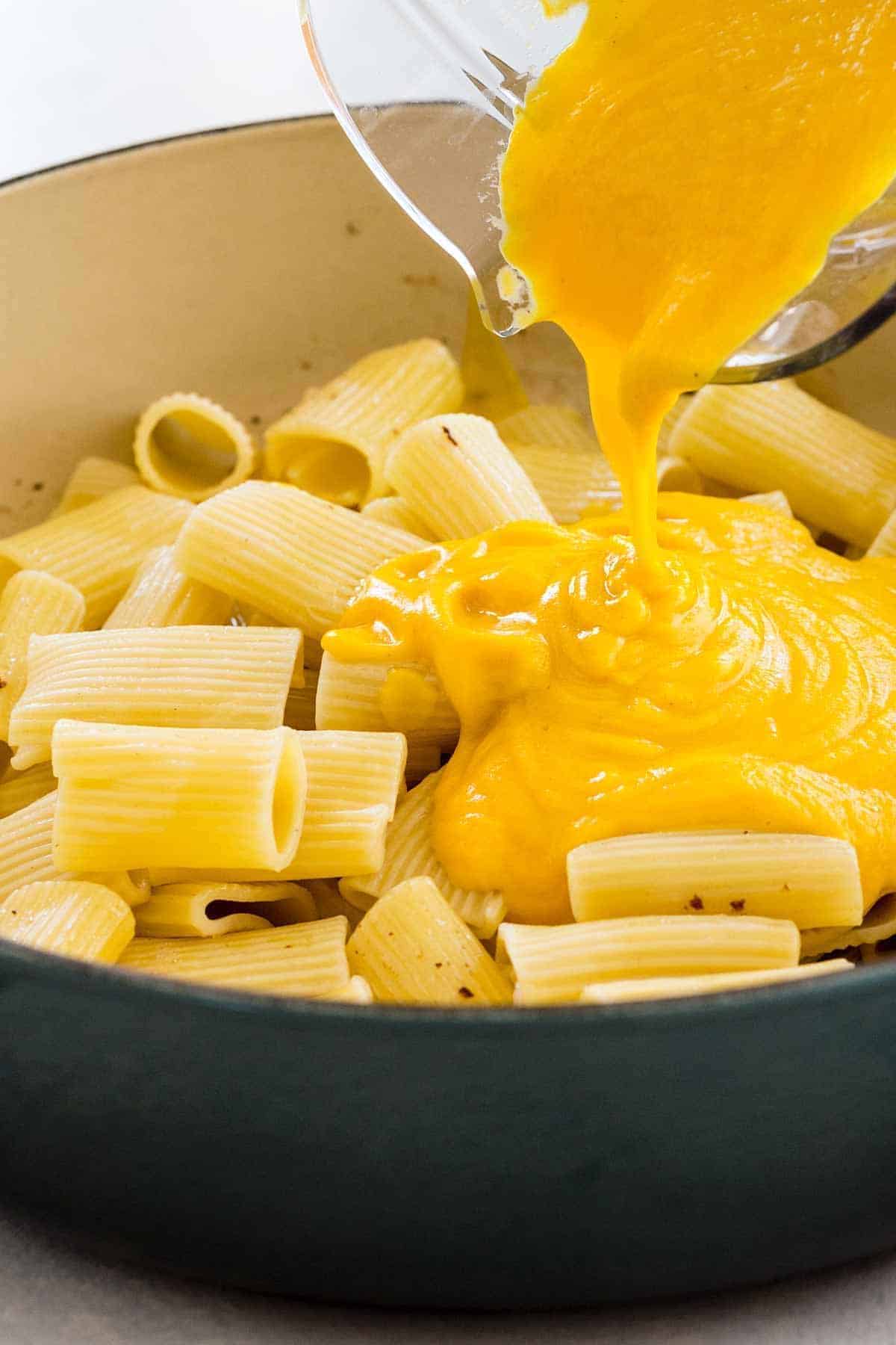 Pouring creamy butternut squash sauce from a blender pitcher over cooked rigatoni pasta in a pot.