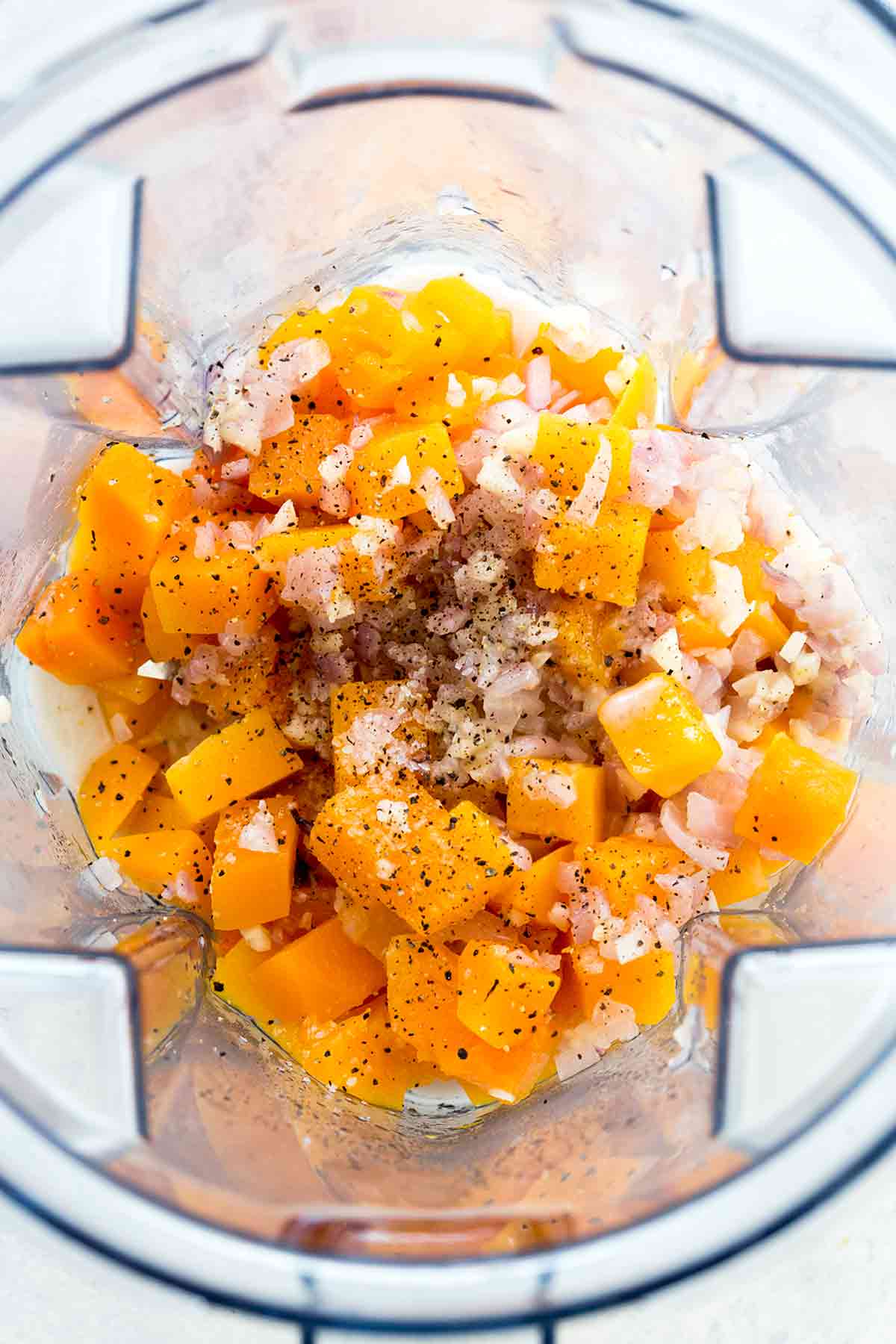 Diced butternut squash, garlic, shallots, and black pepper inside a blender jar before puréeing.