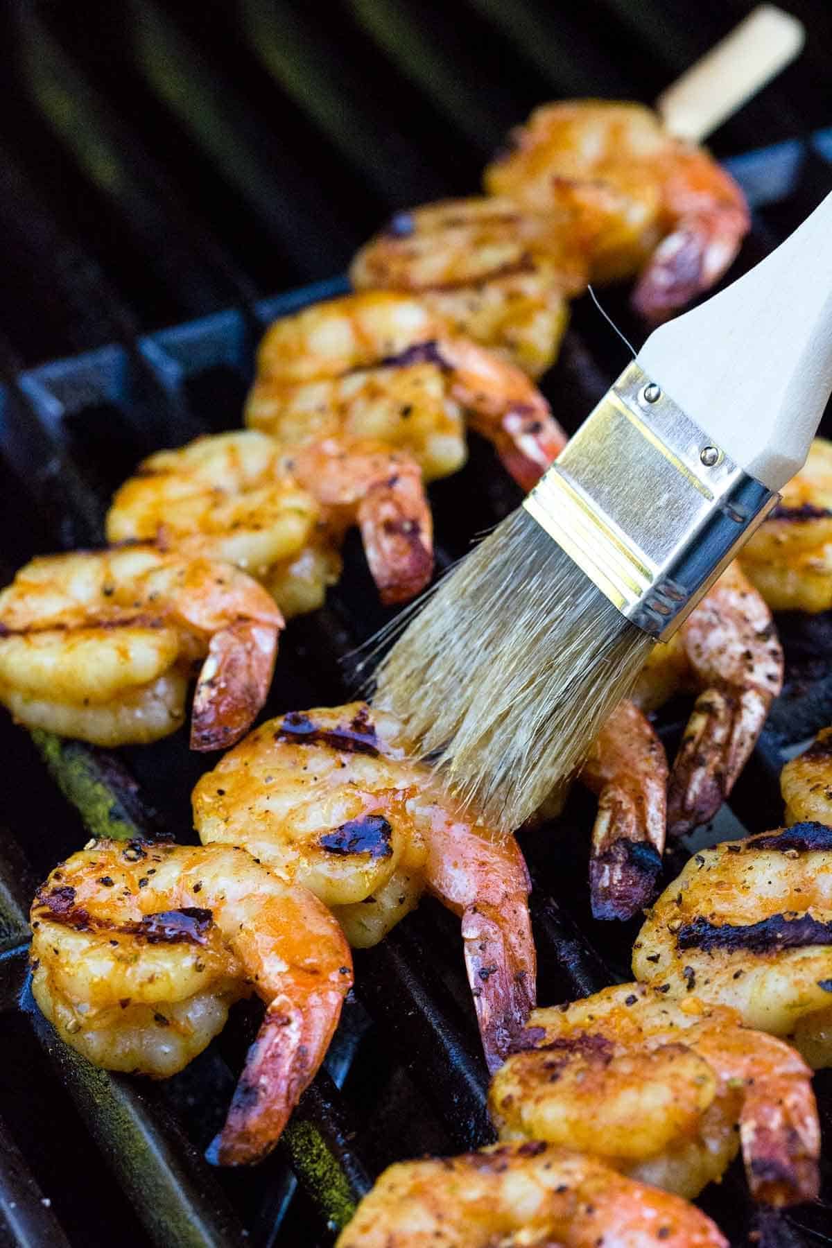 Shrimp skewers on the grill with char marks being basted with butter using a pastry brush.
