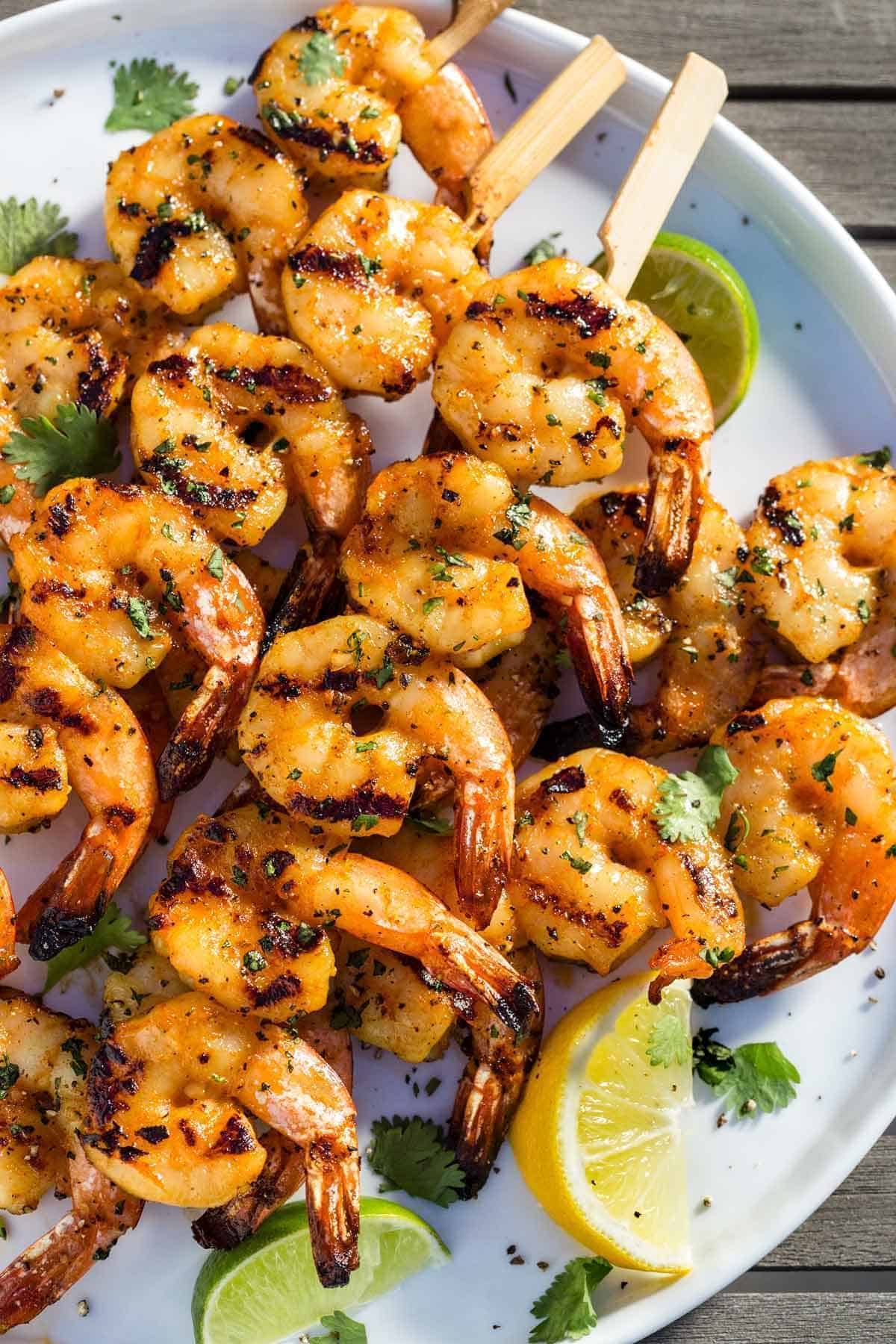 Plate of grilled shrimp skewers with charred grill marks, garnished with cilantro and lemon and lime wedges.