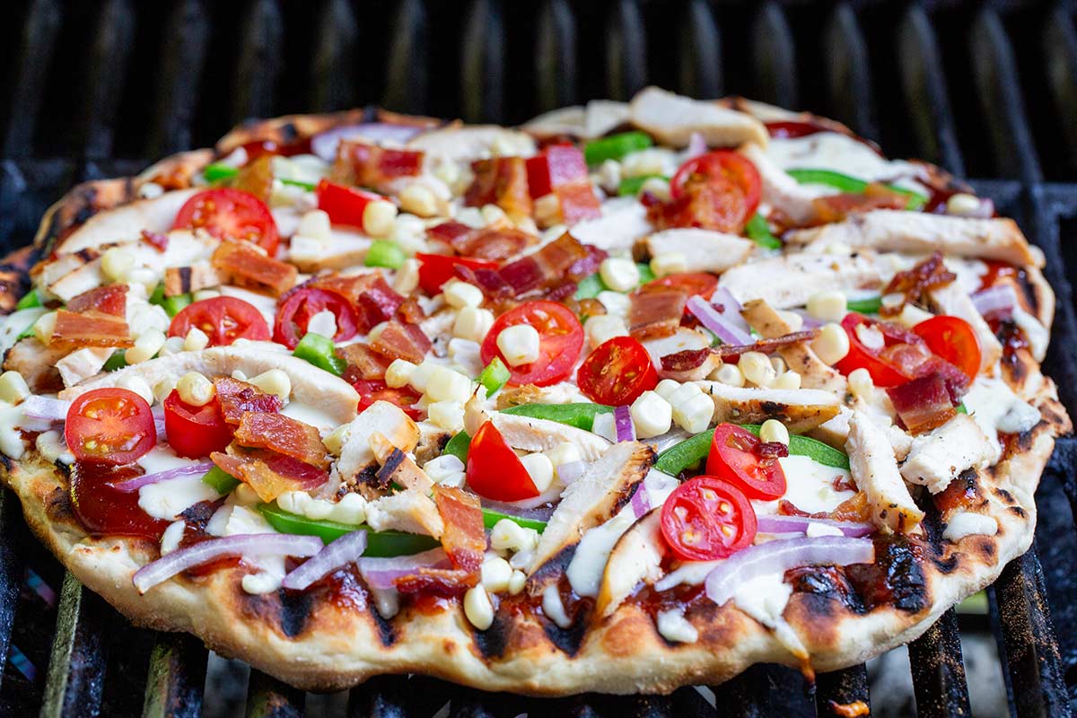 Grilled pizza crust on barbecue grates topped with sliced chicken, bacon, cherry tomatoes, red onion, green peppers, and corn.