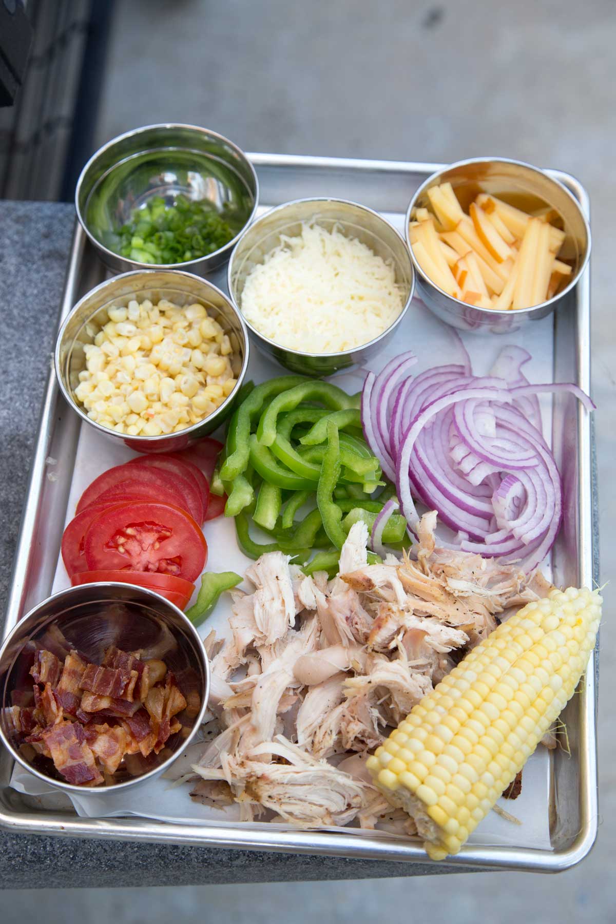 Sheet pan filled with prepped pizza ingredients, including shredded chicken, bacon, sliced tomatoes, green peppers, red onions, corn, and cheeses.