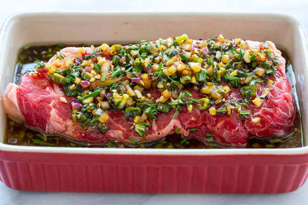 Raw steak in a baking dish topped with chopped herbs, garlic, and marinade.