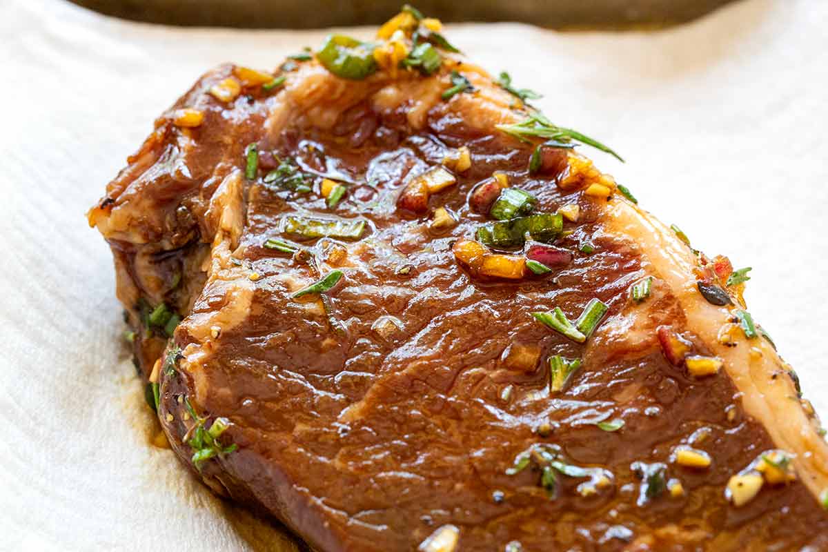 Raw steak coated in liquid marinade with herbs, minced garlic, and chopped green onions.