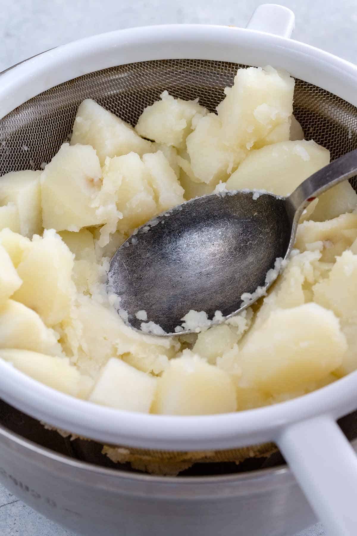 Metal spoon pressing potatoes through a fine-mesh sieve and into a bowl.