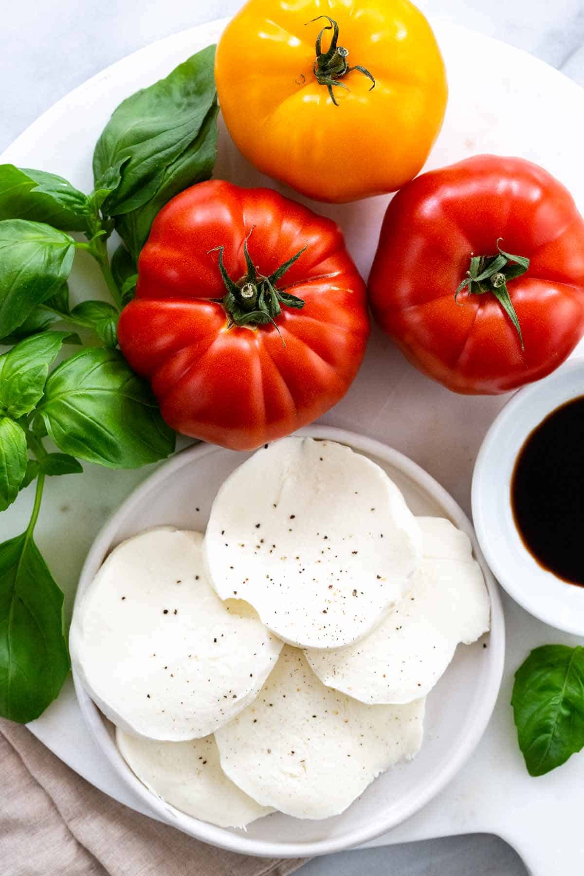 Ingredients for Caprese salad, including red and yellow tomatoes, fresh basil, sliced mozzarella, and balsamic glaze