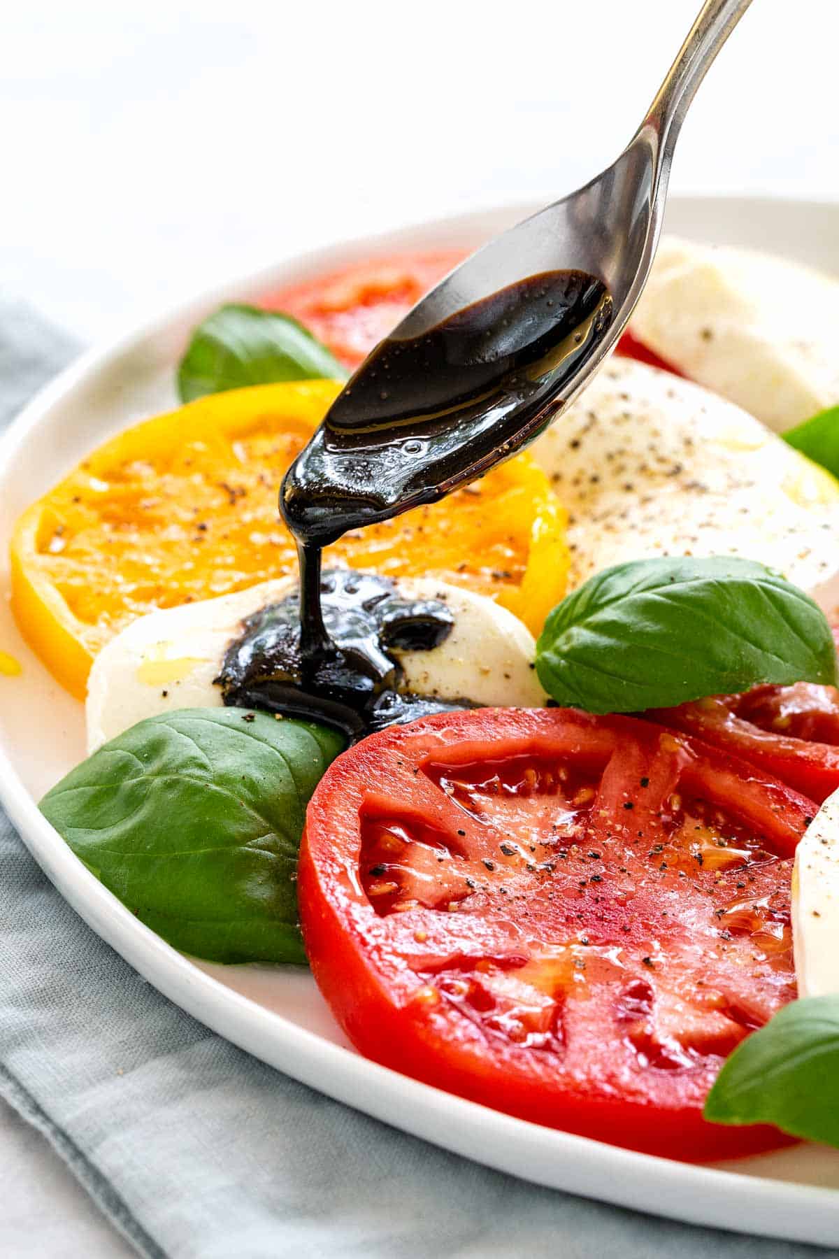 Spoon drizzling thick balsamic reduction over sliced tomatoes, fresh mozzarella, and basil leaves on a platter