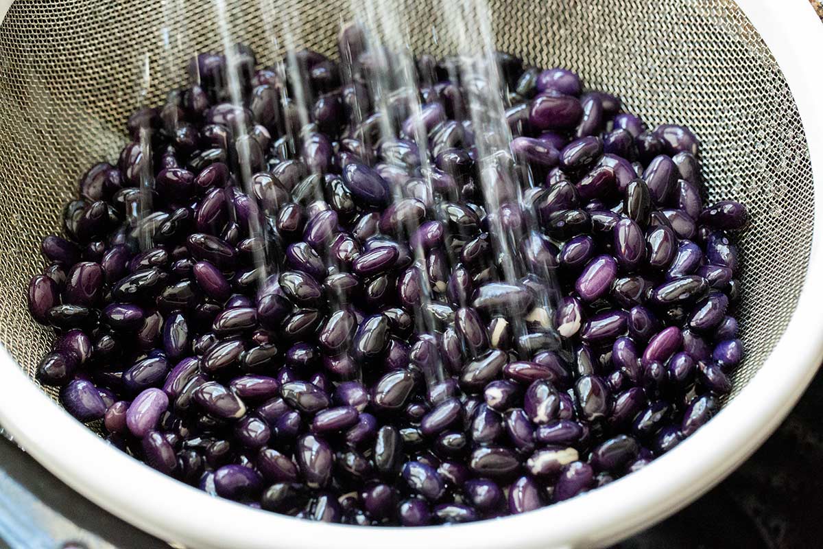 Rinsing dried black beans under running water in a metal mesh colander.