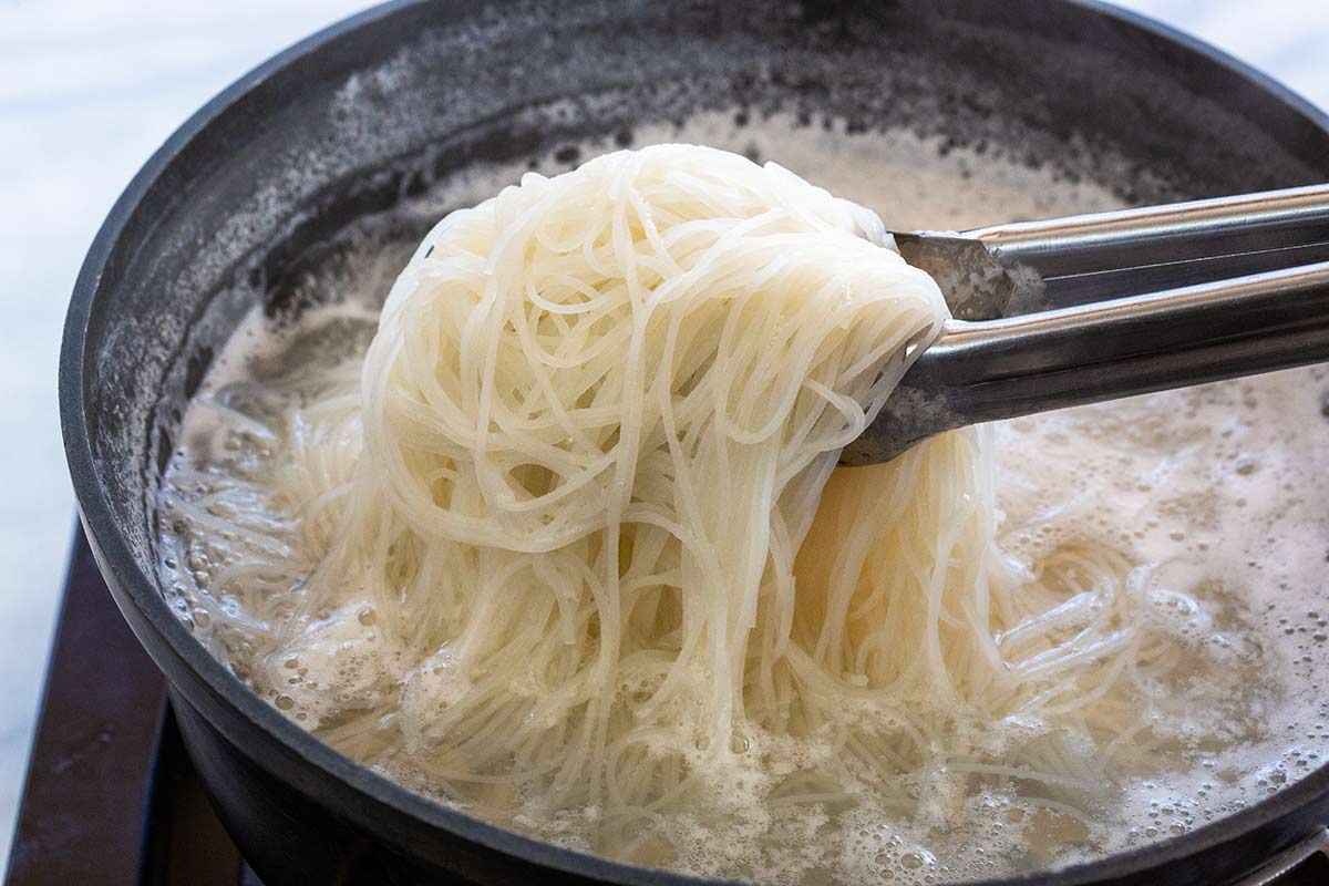 Metal tongs lifting Vermicelli rice noodles cooking in a pot.