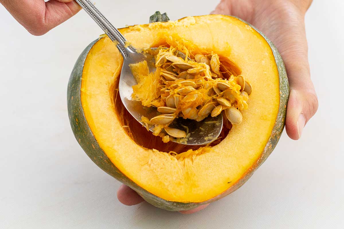 A metal spoon scooping seeds and pulp from the cavity of a halved acorn squash.
