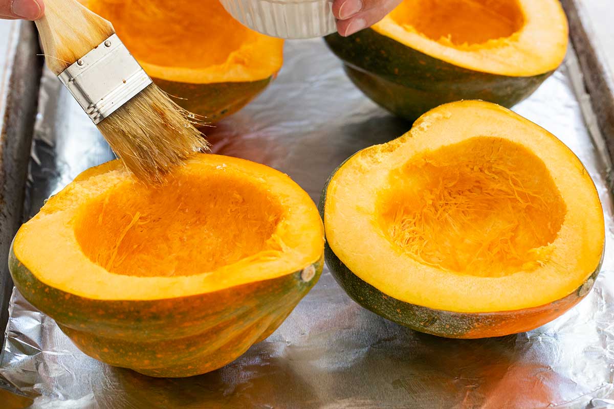 Brushing olive oil onto halved and seeded acorn squash on a foil-lined baking sheet.