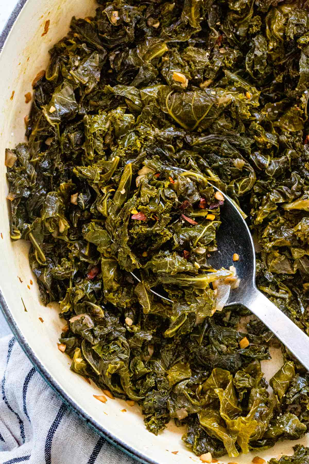 Tender braised kale cooked with garlic and red pepper flakes in a pot, scooped with a metal spoon.