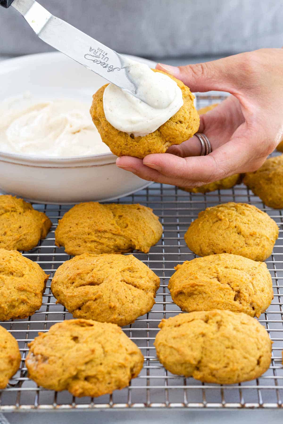 Person spreading cream cheese frosting on top of pumpkin cookies.