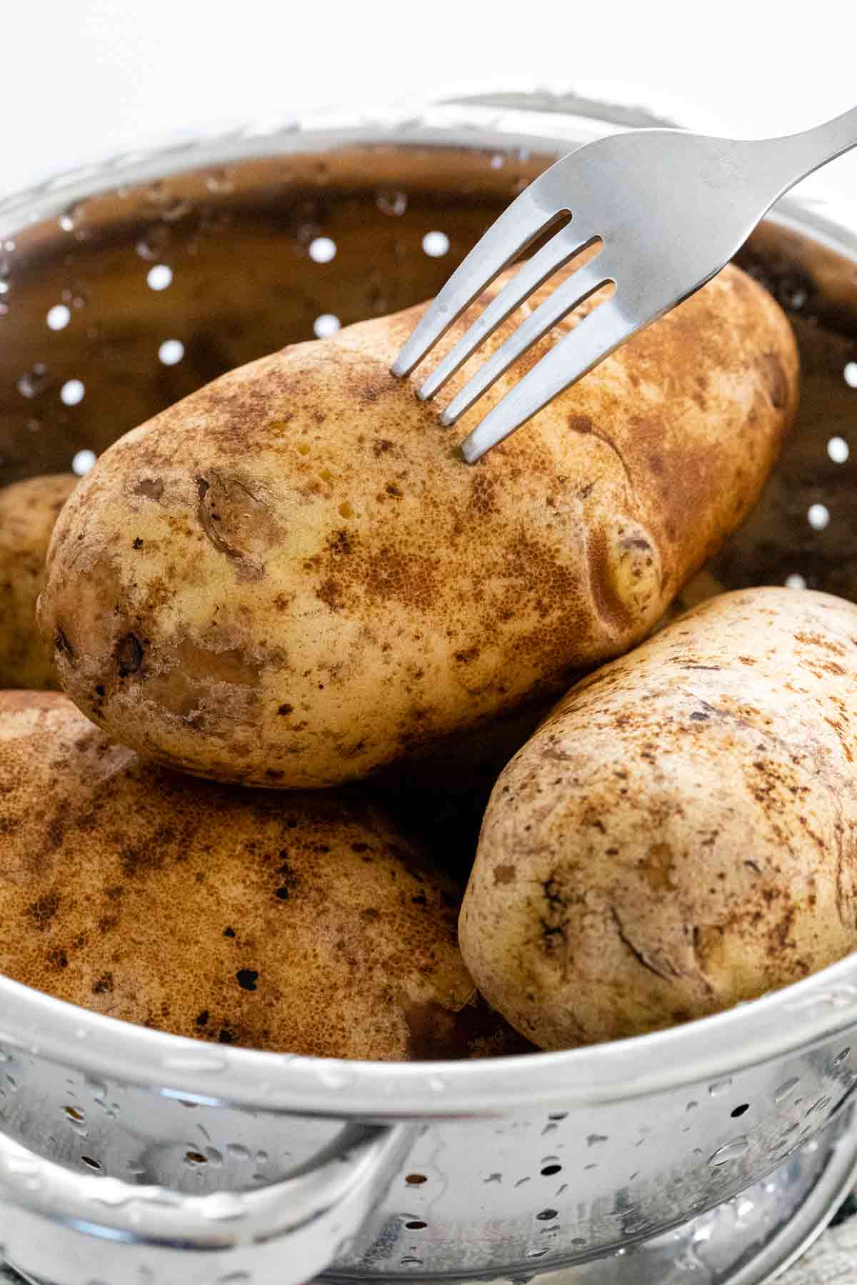 Fork piercing holes into raw potatoes inside a colander.