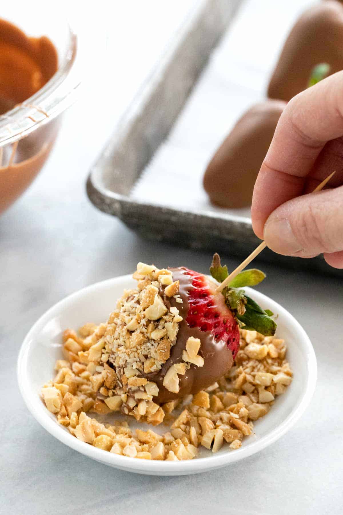 Using a toothpick to hold a chocolate covered strawberry being coated with crushed nuts.