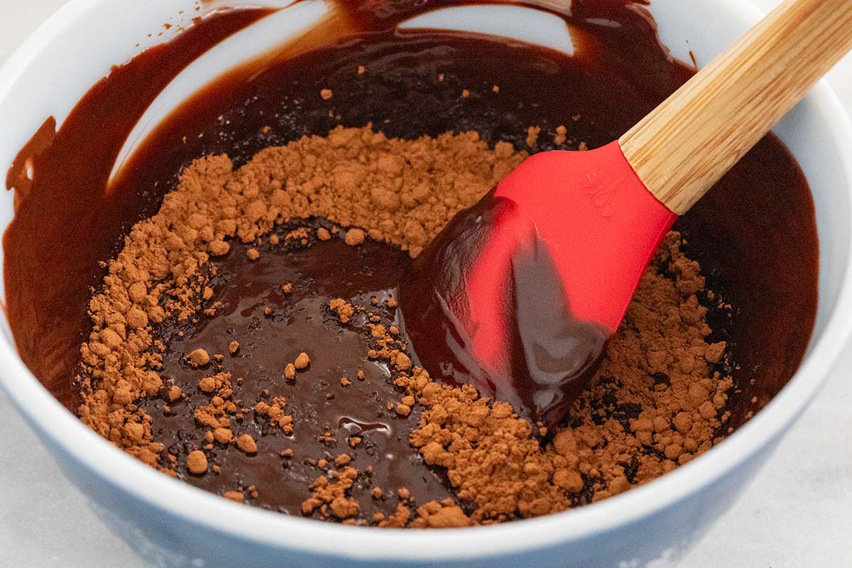 Melted chocolate in a bowl with cocoa powder being mixed in with a spatula.