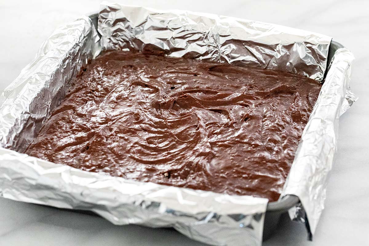 Brownie batter inside a square cake pan lined with foil.