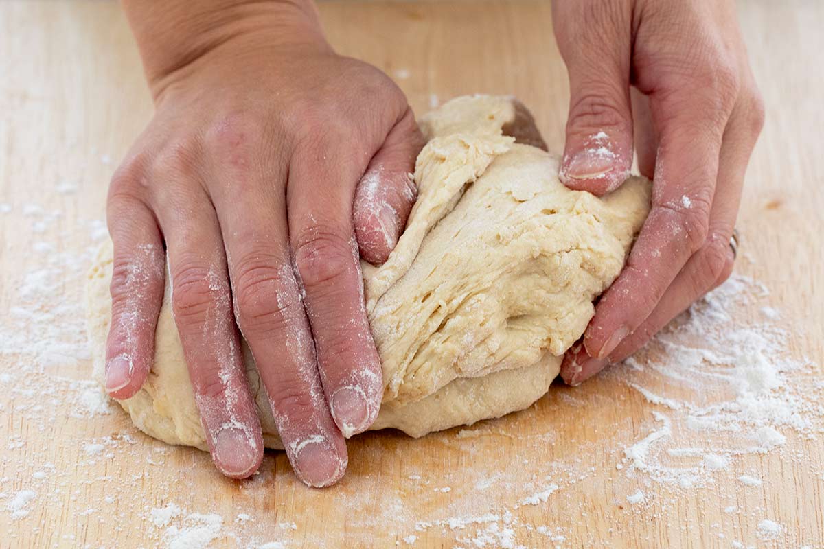 Hands kneading dough on a wooden cutting board dusted with flour.