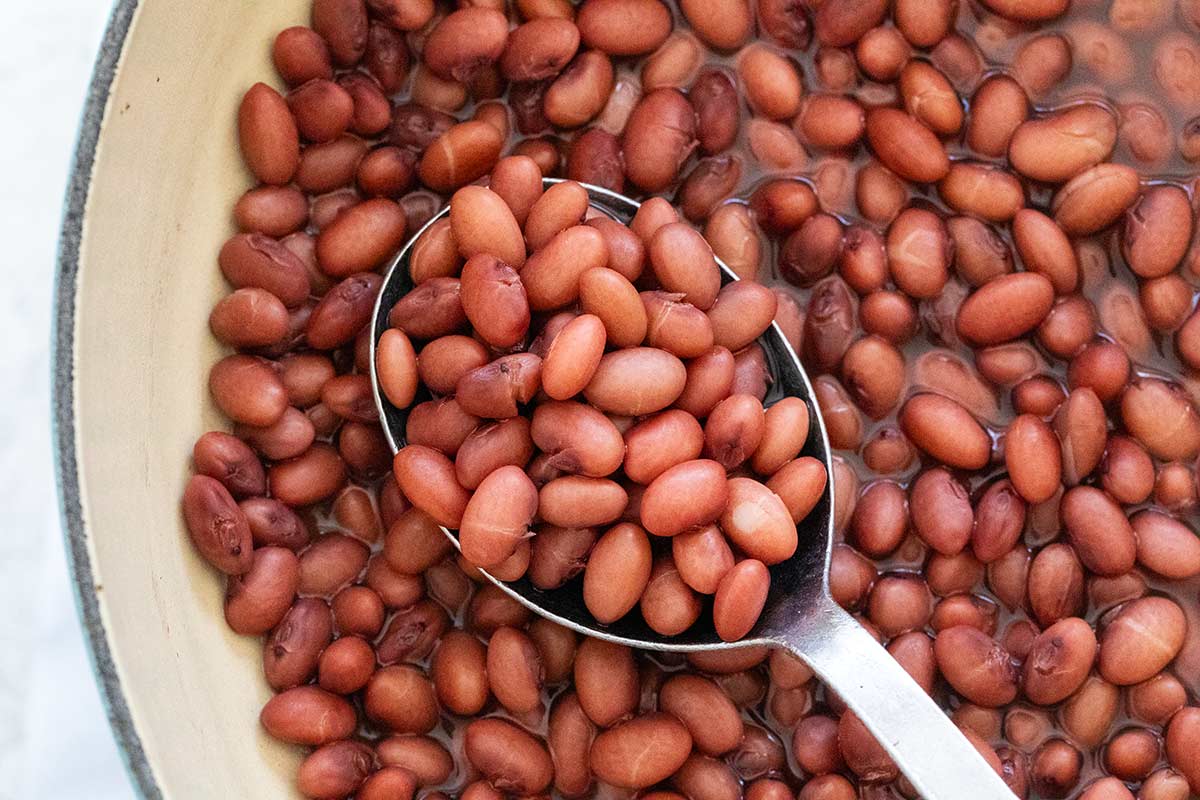 A metal spoon scooping tender cooked beans from a pot.