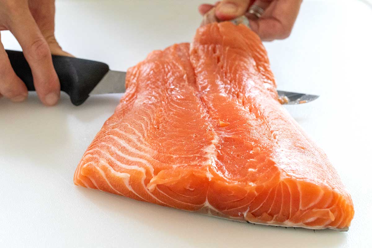 Hands using a knife to slice between the flesh and skin of a raw salmon fillet.
