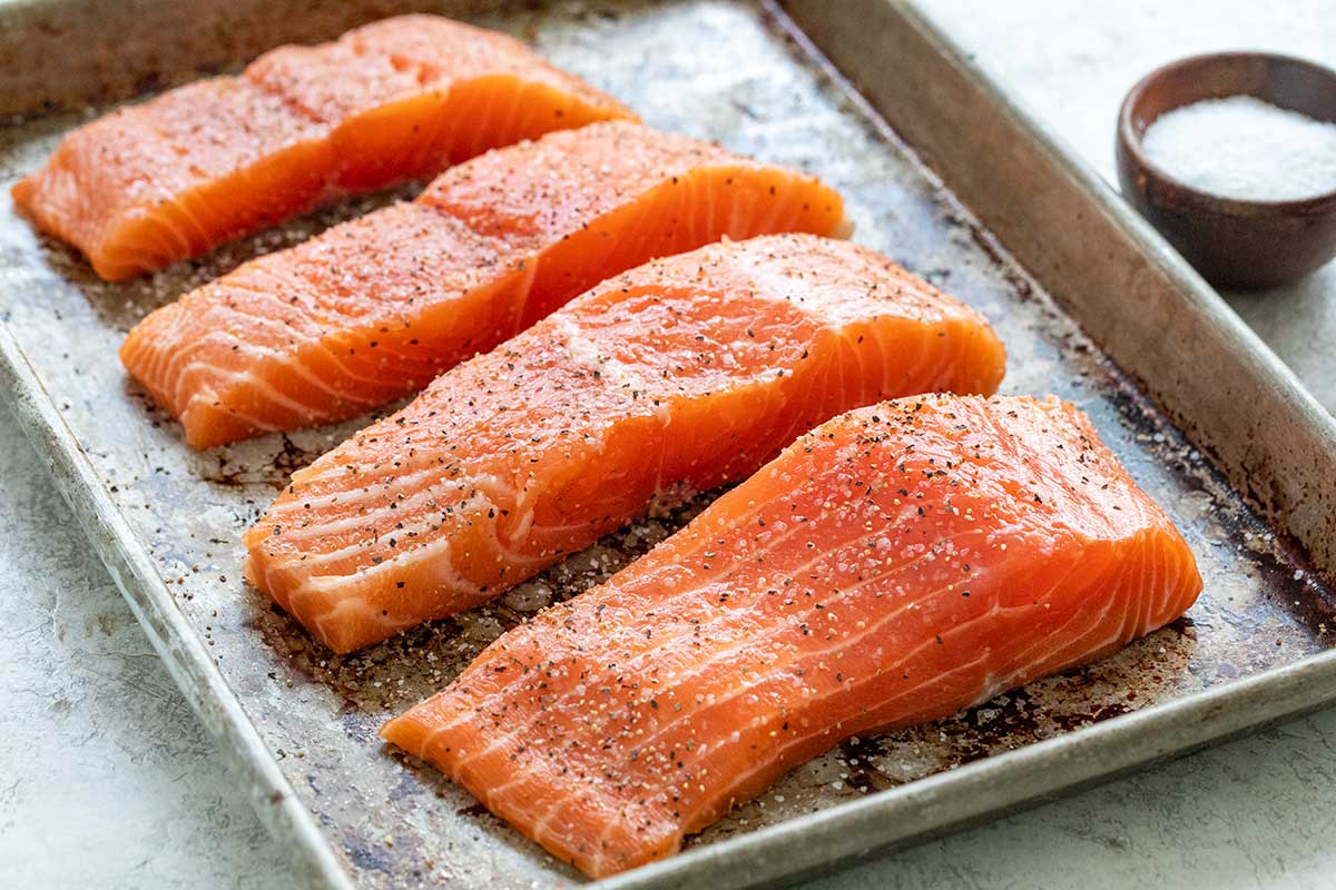 Four raw salmon fillets seasoned with salt and black pepper on a baking sheet.