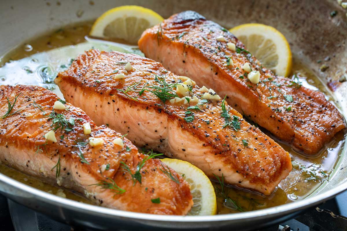 Three pan-seared salmon fillets with golden brown crusts in a skillet with lemon garlic butter sauce and fresh dill.