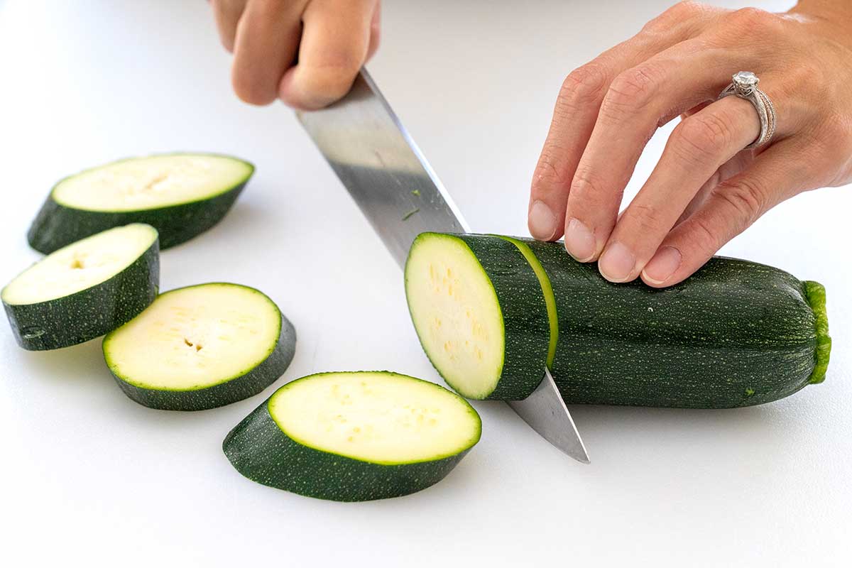 Slicing a fresh zucchini on a bias into thick rounds on a cutting board.