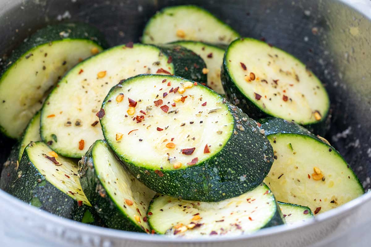 Thick, bias-cut zucchini slices tossed with oil, dried herbs, and red chili flakes in a metal bowl.