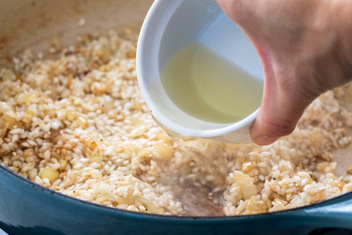 Pouring white wine from a small bowl into toasted arborio rice and onions in a pot.