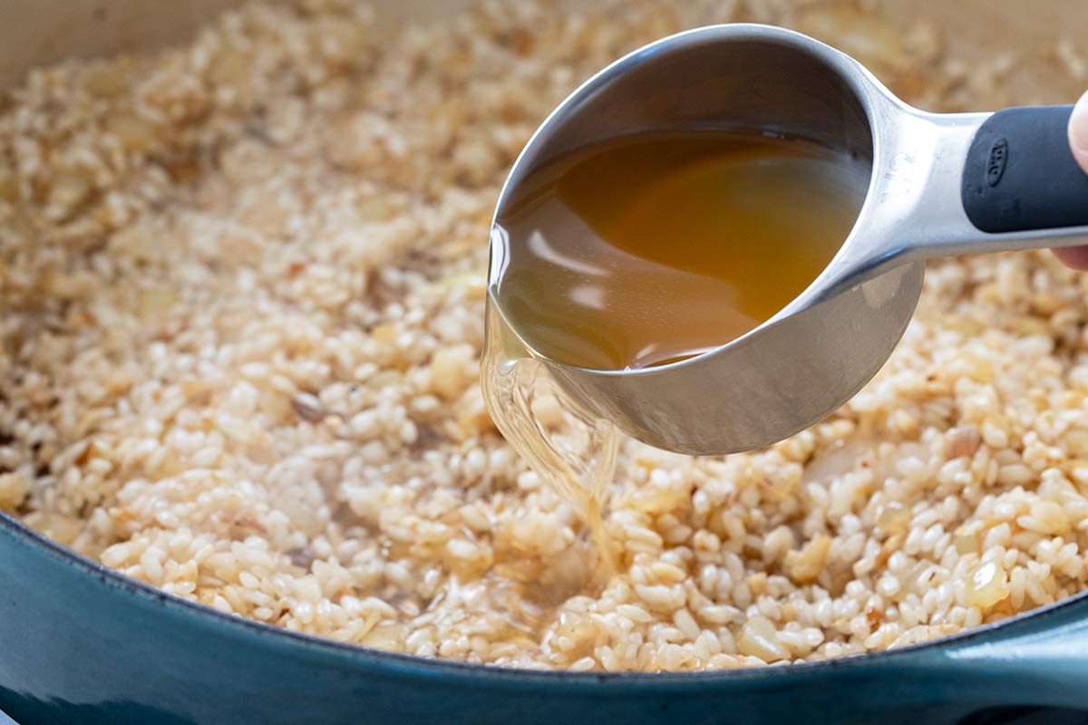 Pouring warm vegetable stock from a measuring cup into a pan of arborio rice.