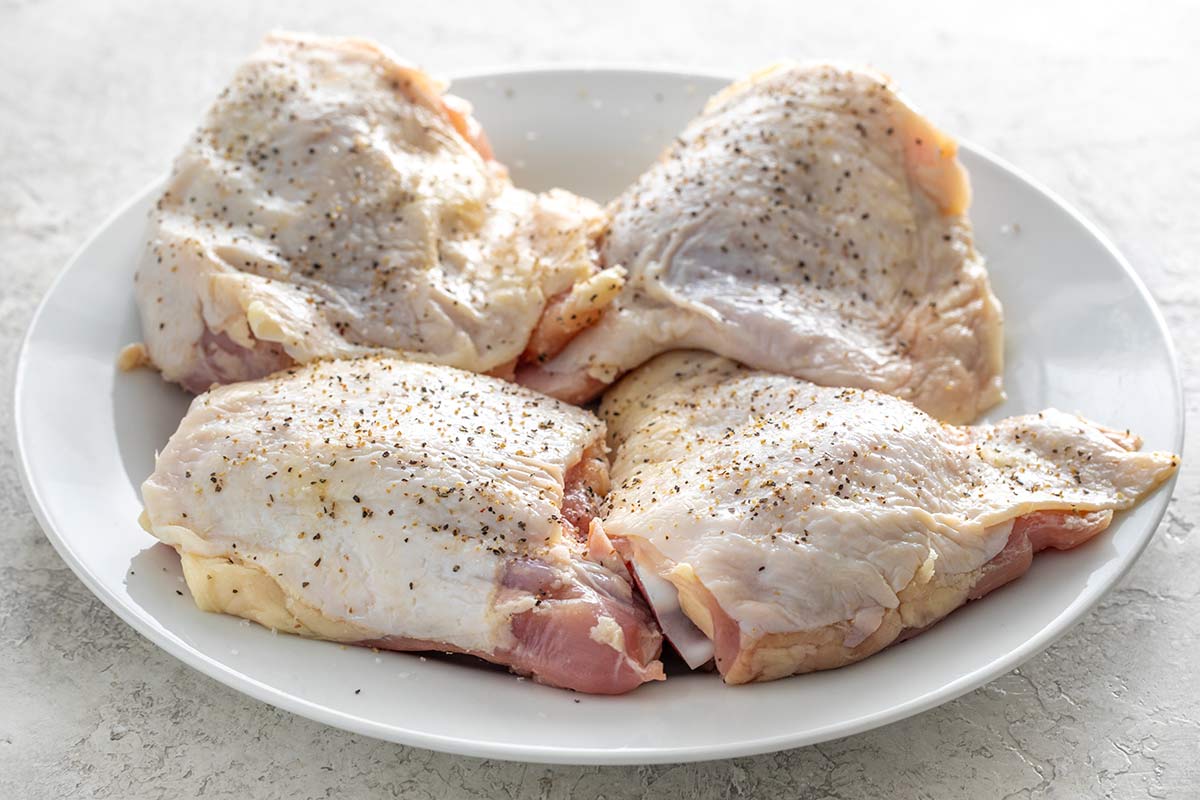 Four raw, skin-on chicken thighs seasoned with salt and black pepper on a white plate.