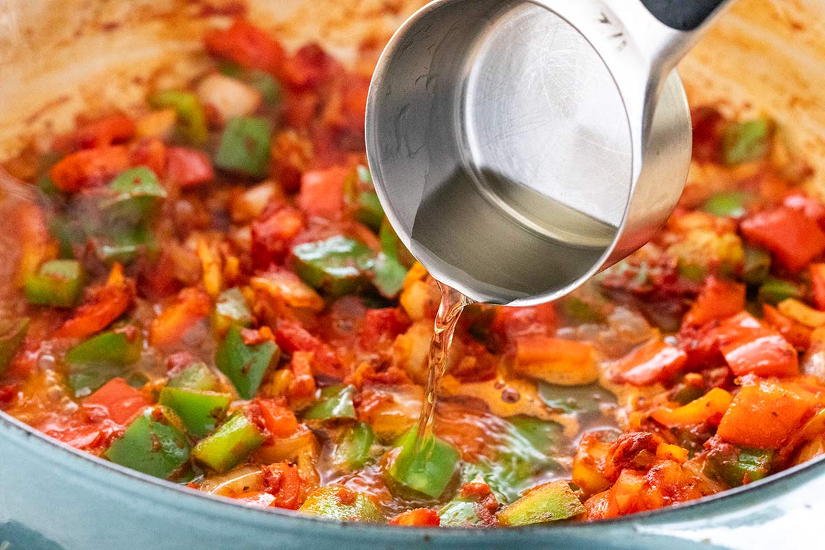 Pouring white wine from a metal measuring cup into sautéed red and green bell peppers in a pot.