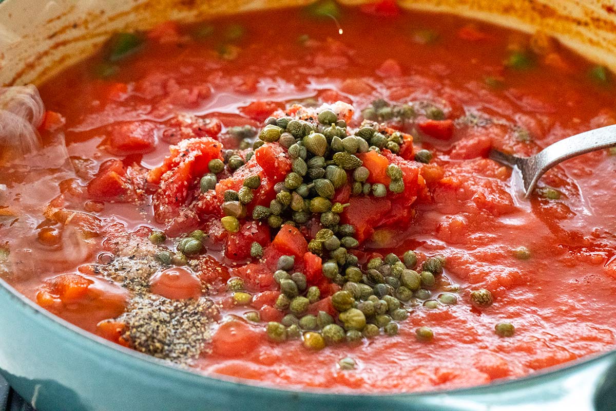 Diced tomatoes, capers, and seasonings added to the tomato sauce base in a braising pot.