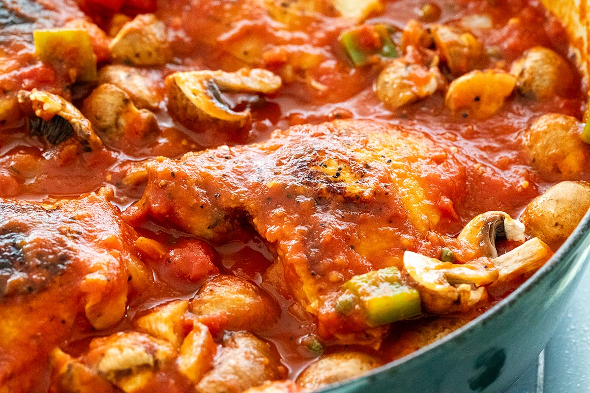 Seared chicken thighs nestled in tomato sauce with mushrooms and bell peppers in a braising pan.