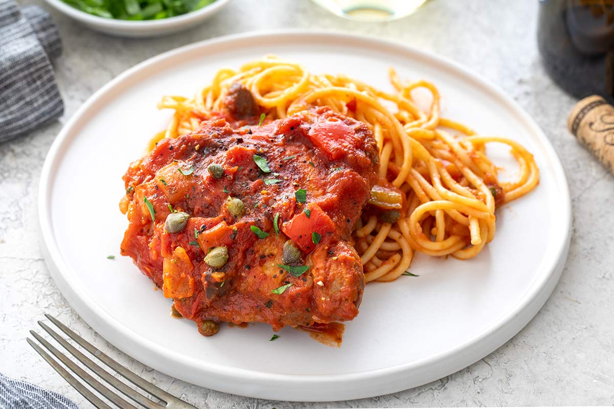 Braised chicken cacciatore thigh topped with tomato sauce and capers, served alongside spaghetti on a white plate.