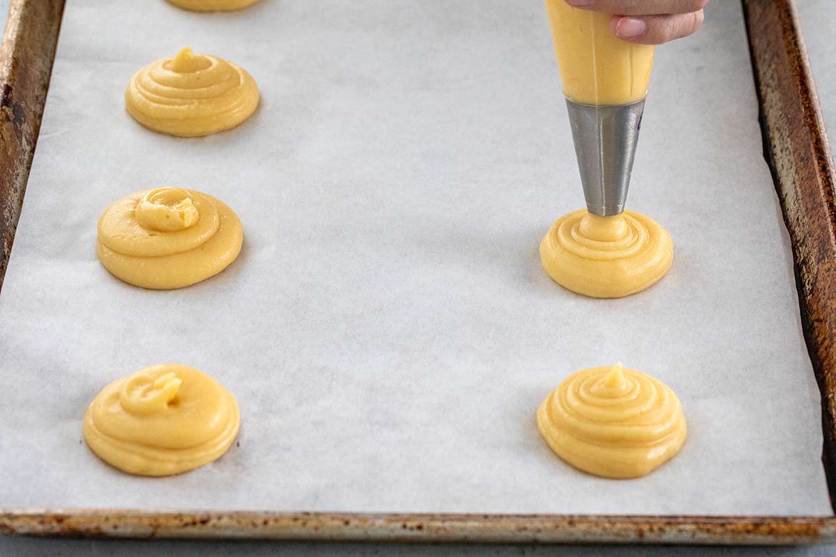Puff pastry being piped into little rounds on a paper lined sheet pan.