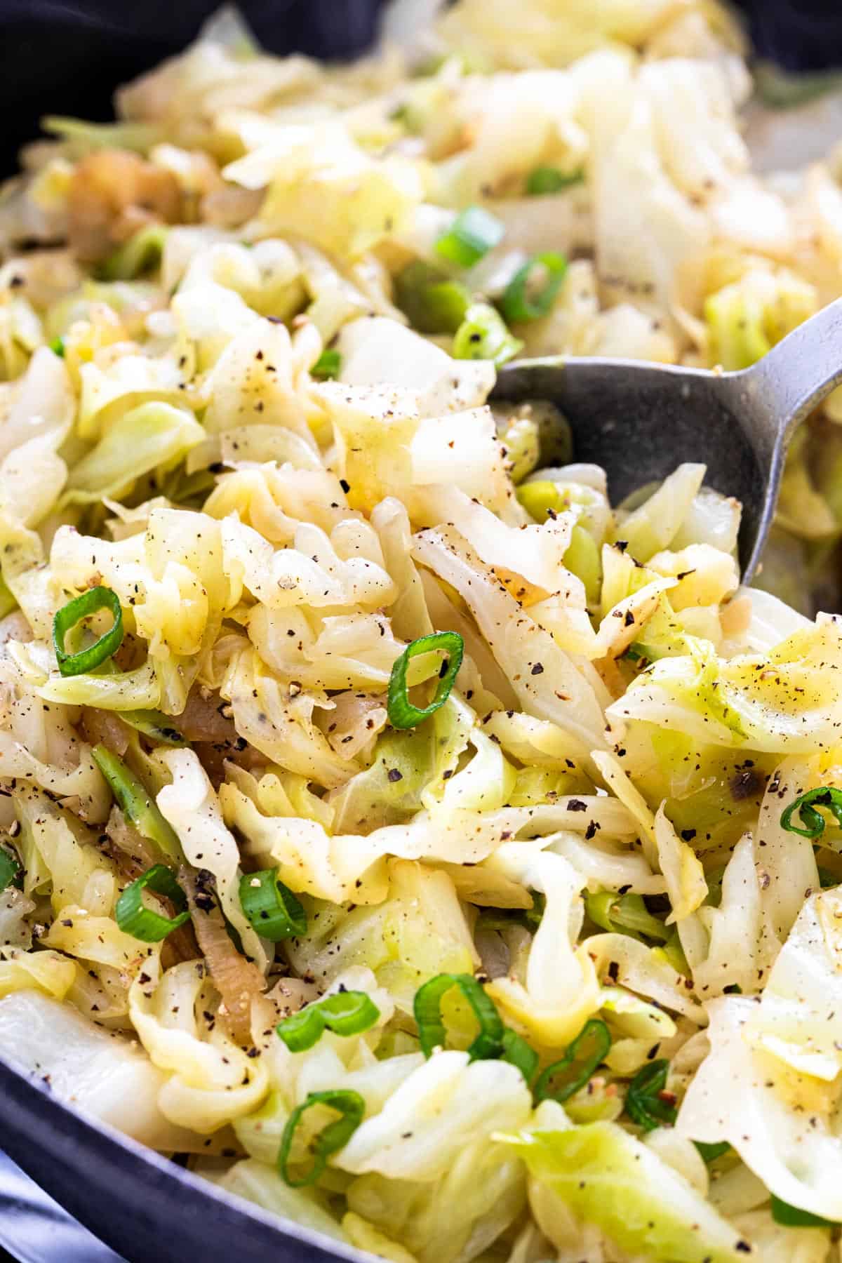 Sautéed cabbage seasoned with black pepper and garnished with sliced green onions in a skillet.
