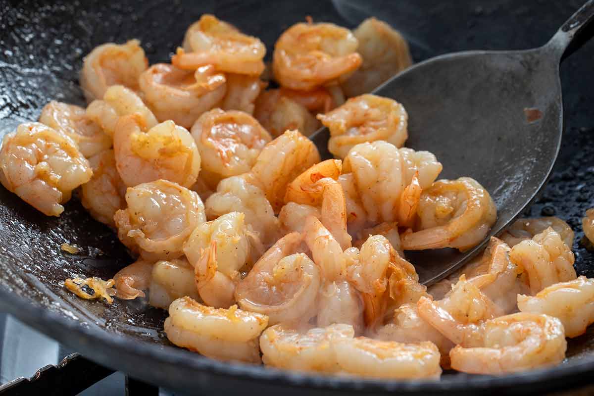 Stir-frying seasoned shrimp in a wok with a metal spatula until pink and opaque