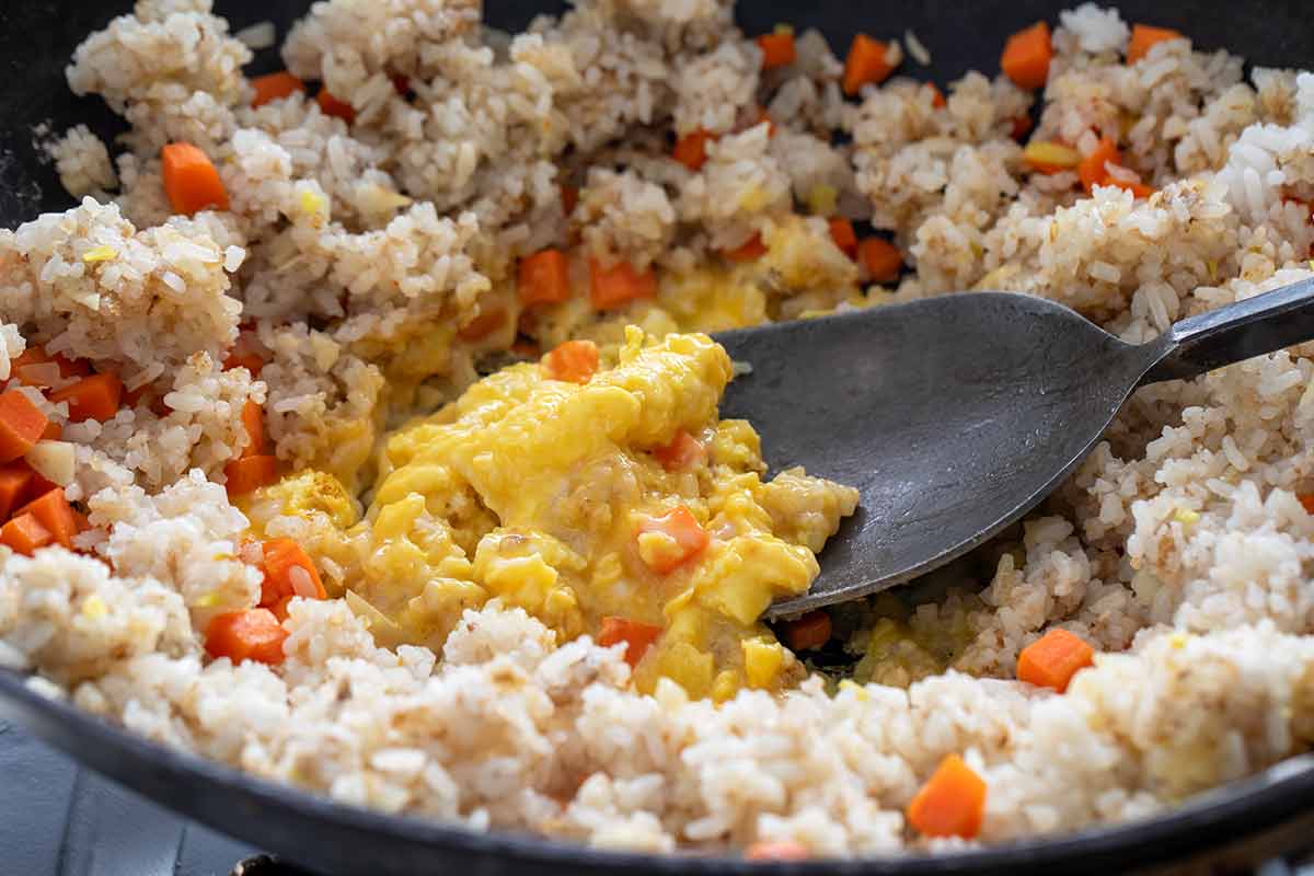 Scrambling beaten eggs in a well in the center of fried rice and diced carrots in a pan